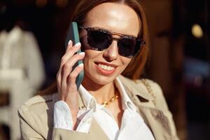 sorridente mulher dentro oculos de sol é falando telefone em cidade rua fundo e olhando às Câmera foto