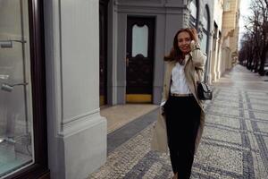 atraente elegante mulher dentro casaco cruzando a cidade rua e olhando às Câmera foto