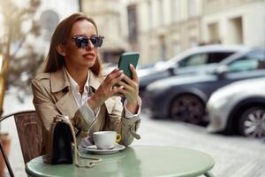 sorridente à moda mulher dentro óculos sentado às cafeteria terraço e segurando telefone enquanto bebendo café foto