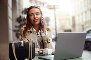 sorridente mulher falando telefone enquanto trabalhando conectados sentado às cafeteria terraço. borrado fundo foto