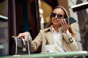 mulher vestindo óculos gravação audio mensagem em Móvel telefone enquanto bebendo café em cafeteria terraço foto