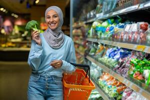 retrato fêmea comprador, muçulmano mulher dentro hijab sorridente e olhando às Câmera, escolhendo legumes e frutas dentro uma ampla supermercado loja, segurando uma cesta com bens dentro mãos, mercearia departamento. foto