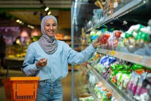 retrato fêmea comprador, muçulmano mulher dentro hijab sorridente e olhando às Câmera, escolhendo legumes e frutas dentro uma ampla supermercado loja, segurando uma cesta com bens dentro mãos, mercearia departamento. foto