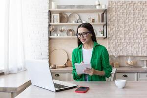 uma jovem lindo mulher é tendo uma controlo remoto trabalho entrevista on-line. sentado às casa dentro a cozinha com uma computador portátil. fala através uma chamar, apresenta ele mesmo, passes a exame, escreve dentro uma caderno. foto