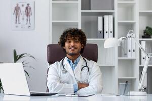 retrato do uma bem sucedido hindu médico dentro a escritório do uma moderno clínica, sentado às a local de trabalho às a mesa sorridente e olhando às a Câmera com dele braços cruzado, uma homem dentro uma branco casaco. foto