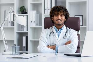 retrato do uma bem sucedido hindu médico dentro a escritório do uma moderno clínica, sentado às a local de trabalho às a mesa sorridente e olhando às a Câmera com dele braços cruzado, uma homem dentro uma branco casaco. foto