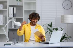 bem sucedido hispânico homem de negocios dentro casa escritório, homem dentro amarelo camisa sentado, recebido carta enviar envelope com notificação mensagem, homem lendo e feliz com conquista, a comemorar vencedor. foto