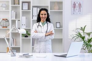 retrato do jovem lindo bem sucedido fêmea médico dentro médico escritório, hispânico mulher com encaracolado cabelo dentro branco médico casaco sorridente e olhando às Câmera com braços cruzado, médico dentro clínica. foto