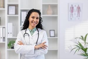 retrato do jovem lindo bem sucedido fêmea médico dentro médico escritório, hispânico mulher com encaracolado cabelo dentro branco médico casaco sorridente e olhando às Câmera com braços cruzado, médico dentro clínica. foto