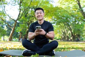 retrato do uma jovem ásia masculino atleta, treinador sentado dentro a parque em uma esteira dentro a lótus posição e usando a telefone. ele parece às a Câmera, sorri. foto