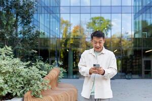 ásia homem usando telefone lado de fora escritório prédio, trabalhador autonomo homem de negocios digitando mensagem e sorridente, programador dentro casual roupas navegando conectados rede Páginas a partir de aplicativo. foto