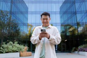 ásia homem usando telefone lado de fora escritório prédio, trabalhador autonomo homem de negocios digitando mensagem e sorridente, programador dentro casual roupas navegando conectados rede Páginas a partir de aplicativo. foto