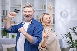 feliz Senior casal dançando dentro seus acolhedor vivo sala, sorridente olhando às Câmera, feliz grandes casado, marido e esposa gastos final de semana às lar. foto