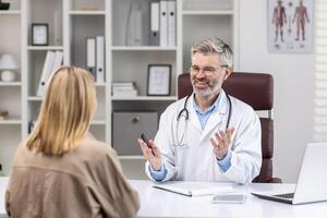 costas visualizar, com experiência maduro médico consultando sorridente fêmea paciente, grisalho homem trabalhando dentro clínica escritório, usando computador portátil. foto