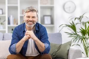 retrato do uma assustador bonito grisalho homem dentro uma azul camisa sentado em a sofá às casa e sorridente às a Câmera, cruzando dele braços dentro frente do ele. foto