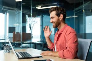 retrato do uma jovem homem dentro uma vermelho camisa. sentado dentro a escritório às a mesa, trabalhando em uma computador portátil, comunicando através da chamar, acenando, sorridente. foto