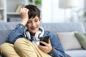 uma sorridente criança do uma Adolescência Garoto dentro fones de ouvido sentado em a sofá às lar, em repouso e usando a telefone. escuta para música, relógios vídeos. ele bate-papos, senta em social redes, tocam conectados jogos. foto