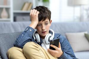 jovem adolescente Garoto lendo mau notícia em telefone, filho sentado em sofá dentro vivo quarto com fones de ouvido às casa fechar-se. foto