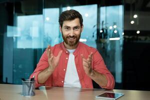 uma jovem masculino professor ensina on-line, condutas palestras, aulas. sentado dentro a escritório às a mesa dentro frente do a Câmera. fala em uma chamar, explica, sorri. foto