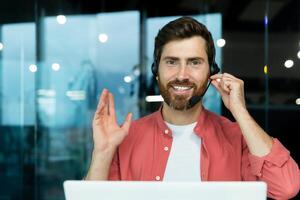 fechar-se retrato do uma jovem homem dentro uma vermelho camisa e uma fone de ouvido trabalhando dentro a escritório em uma computador portátil on-line, dizendo Olá e sorridente às a Câmera. foto