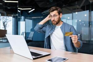 falência, financeiro problemas, mal sucedido conectados compras. chocado jovem homem sentado dentro a escritório, segurando uma crédito cartão, olhando às uma computador portátil. Verificações a conta, arranhões a costas do a cabeça. foto