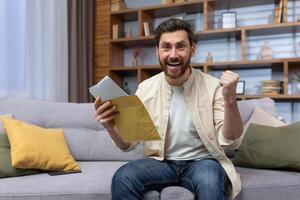 retrato do feliz homem às casa sentado em sofá alegria e a comemorar vitória e sucesso olhando às Câmera e segurando mão acima gesto do triunfo, homem recebido envelope enviar carta com Boa notícias. foto