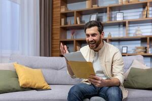 uma alegre jovem homem é sentado em a sofá às casa e segurando a envelope com uma carta dentro dele mãos. recebido Boa notícias, feliz. foto