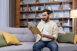 chateado jovem homem sentado em sofá às lar, recebido uma carta, lê mau notícias. divórcio documentos, financeiro dívida, aluguel, crédito, médico resultados. foto