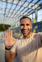 uma jovem africano americano masculino atleta é em pé dentro uma estádio, segurando uma telefone e falando em uma ligar através fones de ouvido. ele parece às a Câmera, sorrisos, diz olá. vertical, fechar acima foto. foto