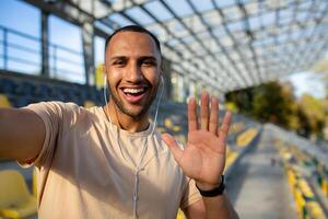 desportista corredor dentro estádio levando selfie foto e falando em ligar com amigos, hispânico homem olhando às Câmera e sorridente gravação Esportes blog, jovem homem depois de ativo treinamento.