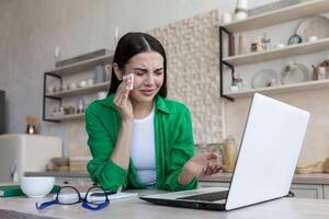 uma jovem mulher senta às casa dentro a cozinha dentro frente do uma computador portátil e chora, lenços dela lágrimas com uma guardanapo, pontos dela mão às a tela, comunica em uma chamar, lê a notícias, recebido mau notícias. foto