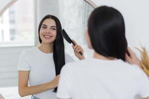 anúncio, cabelo Cuidado. sorridente jovem lindo mulher carrinhos dentro frente do a espelho e pentes dela cabelo com uma escovar. foto