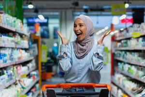 retrato do bem sucedido satisfeito mulher comprador dentro supermercado, alegre muçulmano mulher dentro hijab com compras carrinho entre prateleiras com bens olhando às vários lojas. foto