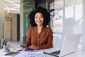 sorridente mulher sentado às uma escrivaninha dentro a escritório segurando uma calculadora, africano americano Gerente às trabalhar, olhando às a Câmera. foto