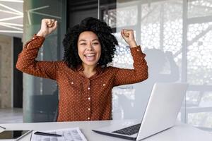 feliz fêmea empresária alegremente levantando dela mãos acima dentro a escritório, africano americano mulher obteve uma promoção, Ganhou a loteria. foto