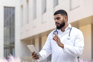 muçulmano jovem masculino médico em pé em a rua perto a hospital e falando em uma ligar a partir de uma tábua, conectados consulta com uma paciente. foto
