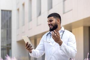 uma jovem muçulmano masculino médico carrinhos perto a hospital em a rua dentro uma branco casaco e fala em a ligar com colegas, uma paciente com uma tábua, consulta, gesticulando com dele mãos. foto