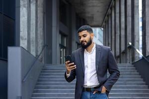 jovem indiano homem, real Estado agente, corretor em pé perto escritório lado de fora e usando telefone, esperando para encontro, digitando mensagem. foto