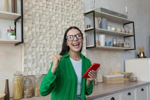 jovem lindo mulher dentro óculos e uma verde camisa às casa dentro a cozinha detém a telefone dentro dela mãos e se alegra. Ganhou a conectados prêmio, loteria, recebido Boa notícias, mensagens. foto