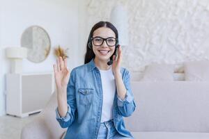 webinário. jovem lindo o negócio mulher dentro óculos e fones de ouvido senta em a sofá às casa e condutas uma webinário, conectados Treinamento em Câmera, consulta, diz, gestos com mãos, sorri. foto