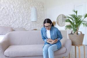 estômago dor. jovem lindo mulher dentro óculos e jeans sentado em a sofá às casa e segurando dela estômago. sente afiado dor, envenenamento, sente mau foto