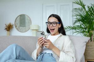 feliz jovem mulher dentro óculos segurando Móvel telefone, a comemorar inesperado vitória dentro jogo, ganho. sentado em sofá às lar, excitado, satisfeito. foto