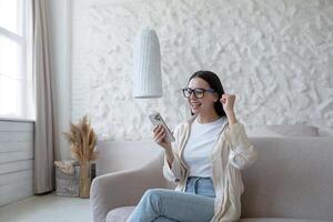 feliz jovem mulher sentado em sofá às casa e segurando telefone. obteve Boa notícias. obteve uma bem sucedido trabalho, passado a entrevista, recebido uma mensagem. ele alegra-se, gritos com alegria, pontos com dele mão sim. foto