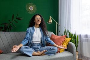 jovem lindo mulher meditando dentro lótus pose, hispânico mulher sorridente contente sonhando sobre futuro planos, sentado em sofá dentro vivo quarto às lar, praticando ioga aula. foto