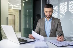 focado homem de negocios analisando papelada dentro uma bem iluminado, contemporâneo escritório configuração com uma computador portátil e moderno Projeto elementos. foto