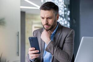 maduro adulto homem de negocios fechar acima pensativamente olhando às telefone tela, homem pensando lendo social meios de comunicação, e-mail, empregado usando aplicativo em Smartphone dentro escritório. foto