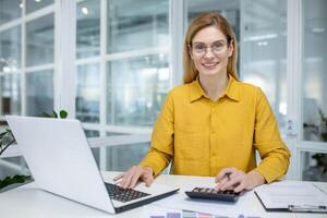 sorridente profissional mulher dentro uma amarelo camisa trabalhando em uma computador portátil e usando uma calculadora dentro uma brilhante moderno escritório meio Ambiente foto