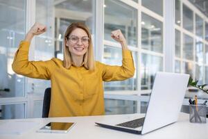 Deleitado empresária levantando dela braços dentro celebração às dela escrivaninha dentro uma moderno escritório, ilustrando carreira sucesso e conquista. foto