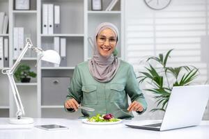 positivo muçulmano mulher sentado às local de trabalho e tendo pausa com salada enquanto trabalhando em sem fio computador portátil. escritório equipado com ajustável luminária e vários suprimentos organizado em prateleiras em fundo. foto