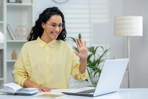 sorridente mulher acenando durante uma ligar em dela computador portátil enquanto sentado às uma branco escrivaninha dentro uma brilhante casa escritório. virtual comunicação, controlo remoto trabalhos dentro uma moderno e acolhedor ambiente. foto